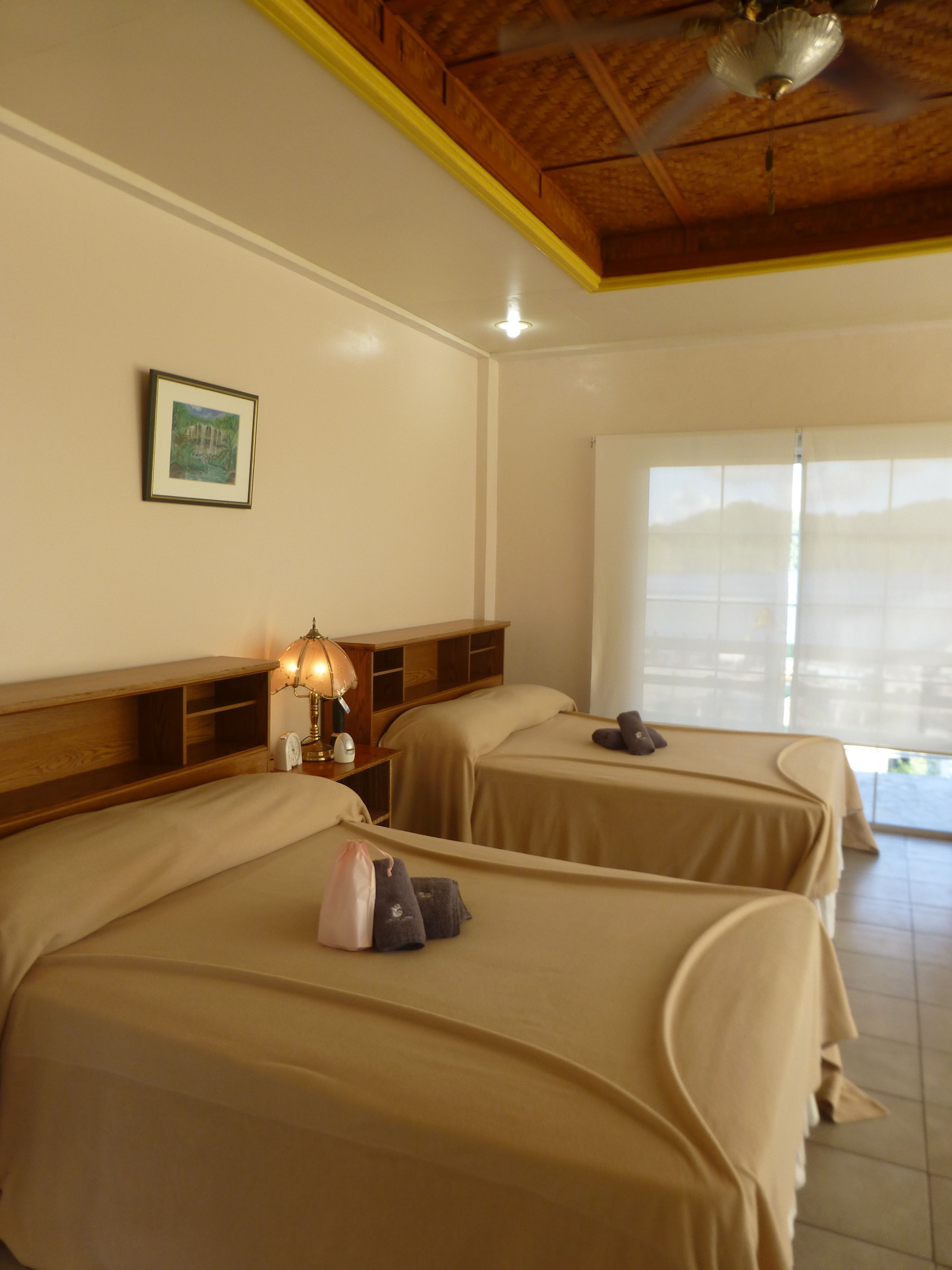 Une chambre d’hôtel avec deux lits, un ventilateur de plafond, une fenêtre avec des stores et un tableau encadré au mur.
