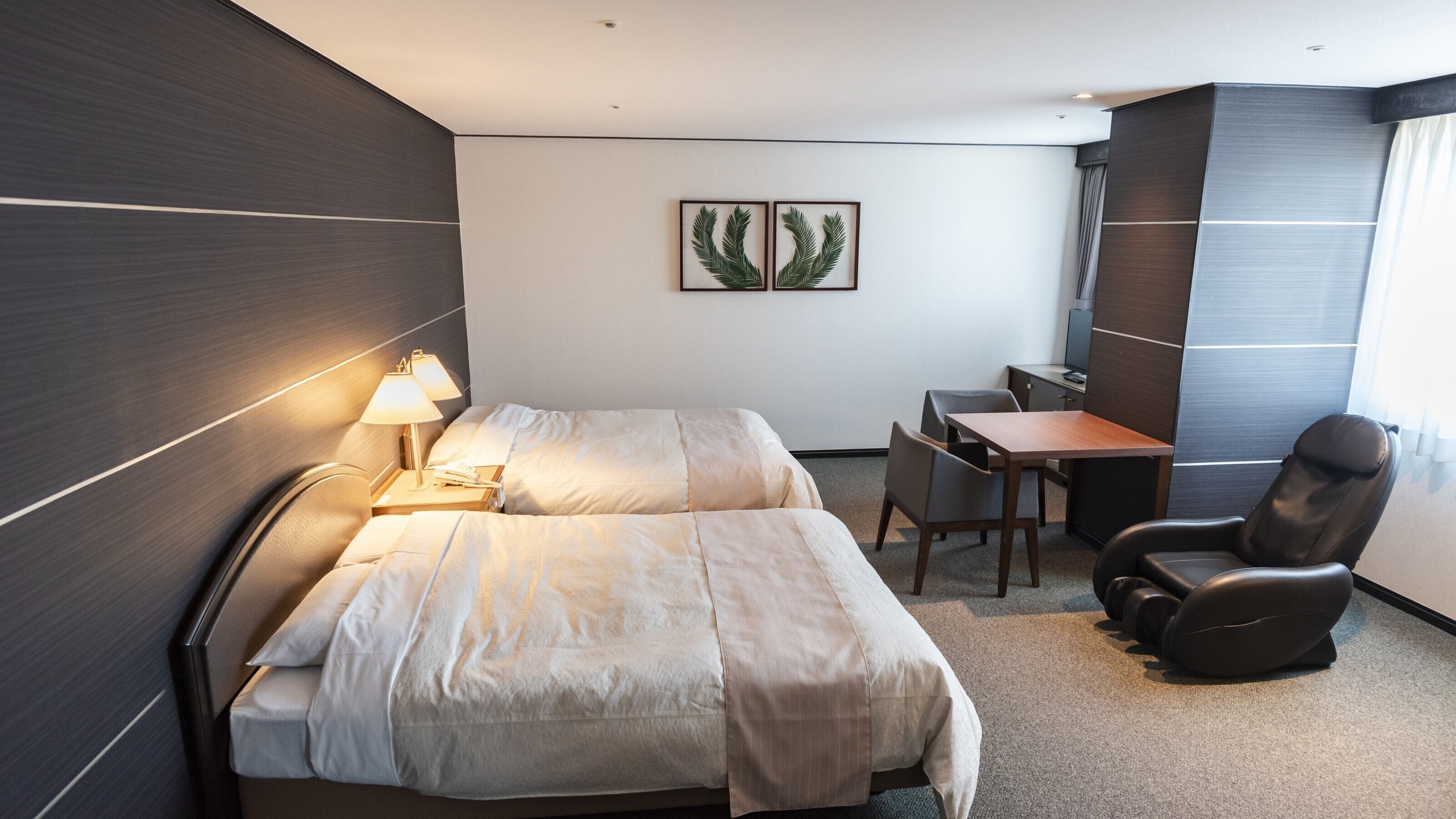 Een hotelkamer met een bed, nachtkastje, lamp, bureau, stoel en een plant in een lijst aan de muur.