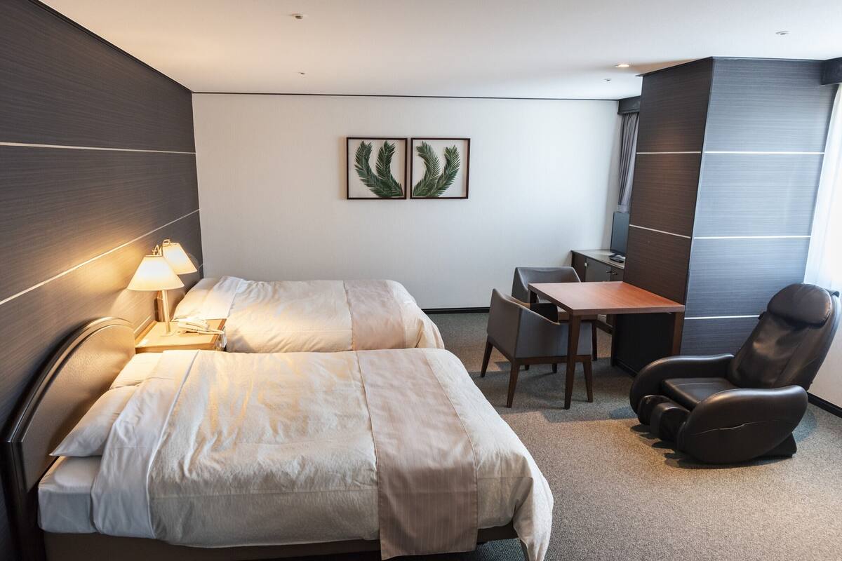 Une chambre d’hôtel comprenant un lit, une table de chevet, une lampe, un bureau, une chaise et une plante décorative accrochée au mur.