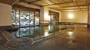Een traditioneel Japans onsen met houten plafond, stenen bassin en uitzicht op een rotsachtig buitengebied.