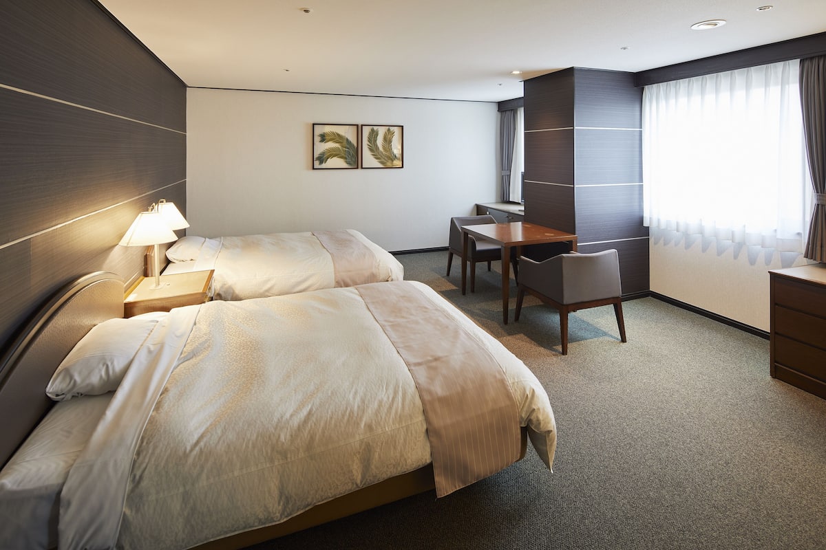 Une chambre d’hôtel avec deux lits, un bureau, une chaise et une grande fenêtre avec des rideaux.