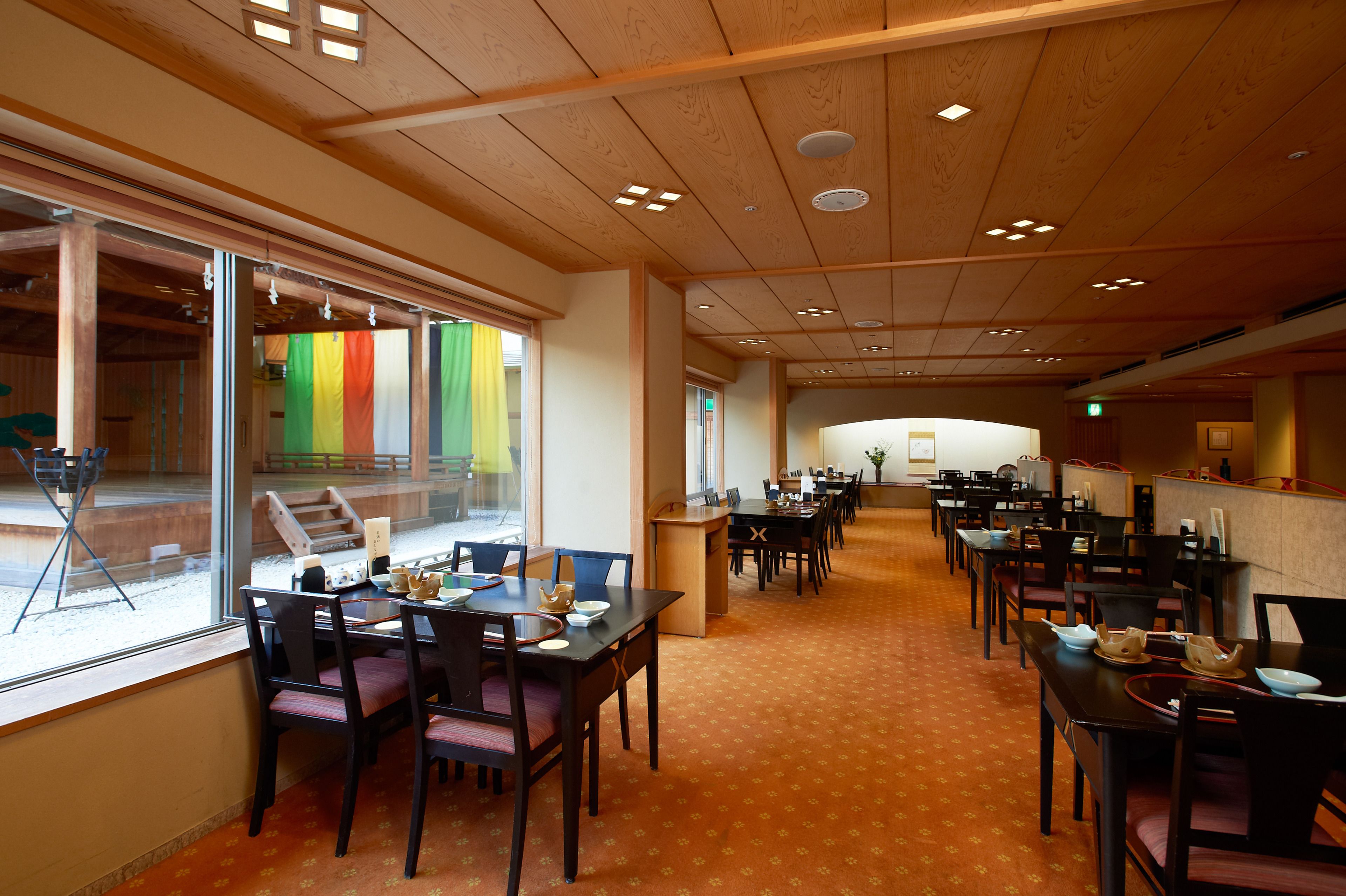 A spacious dining area with multiple tables and chairs, large windows, and a wooden ceiling.