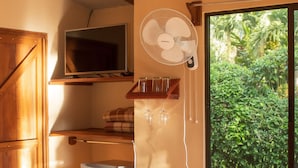 A compact kitchen with a built-in refrigerator, a wooden bench, a wall-mounted fan, and a window with a view of greenery. - Paraiso Volcano Lodge (Guayabo)