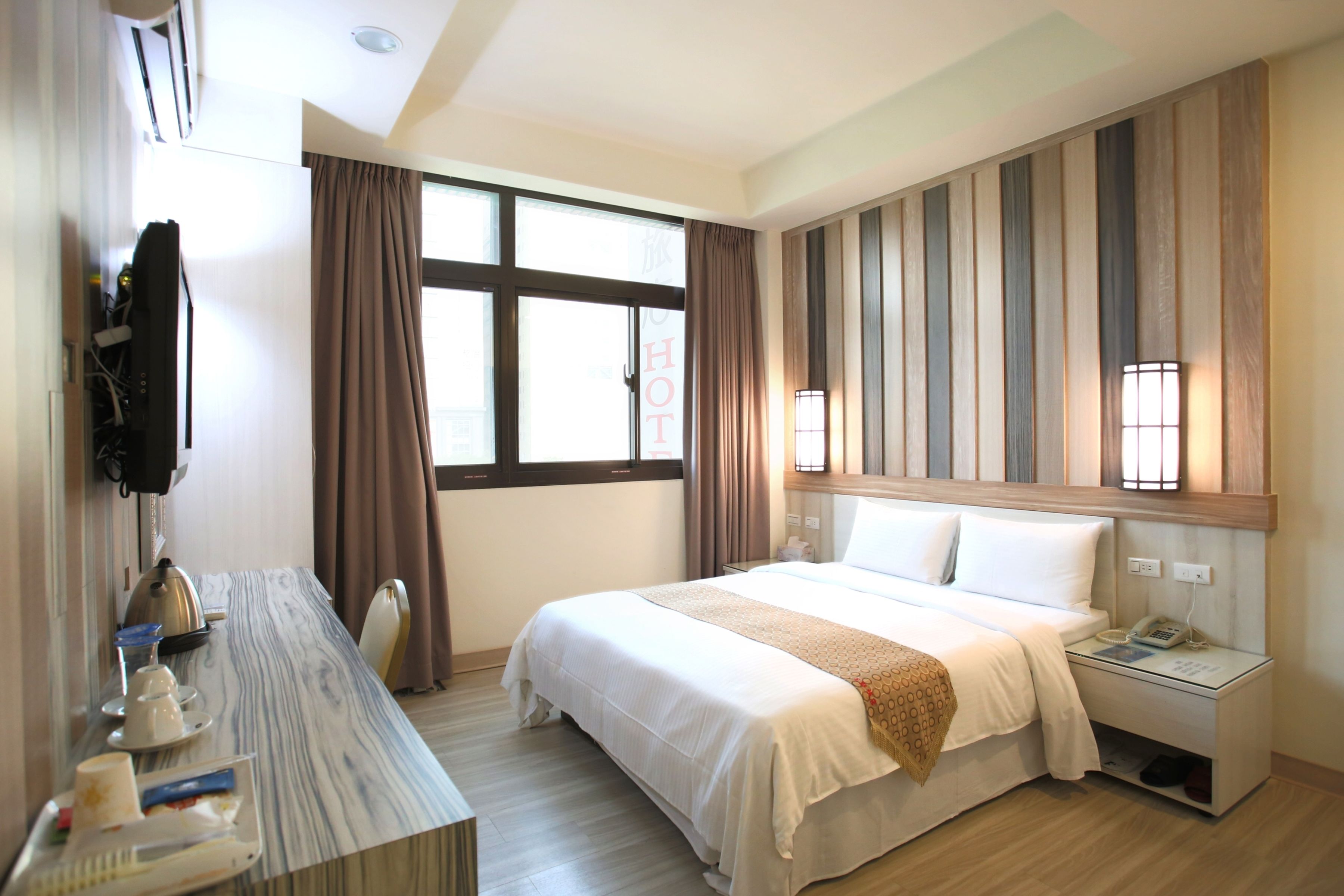 A hotel room with a bed, bedside table, and a desk with a kettle and tea set.