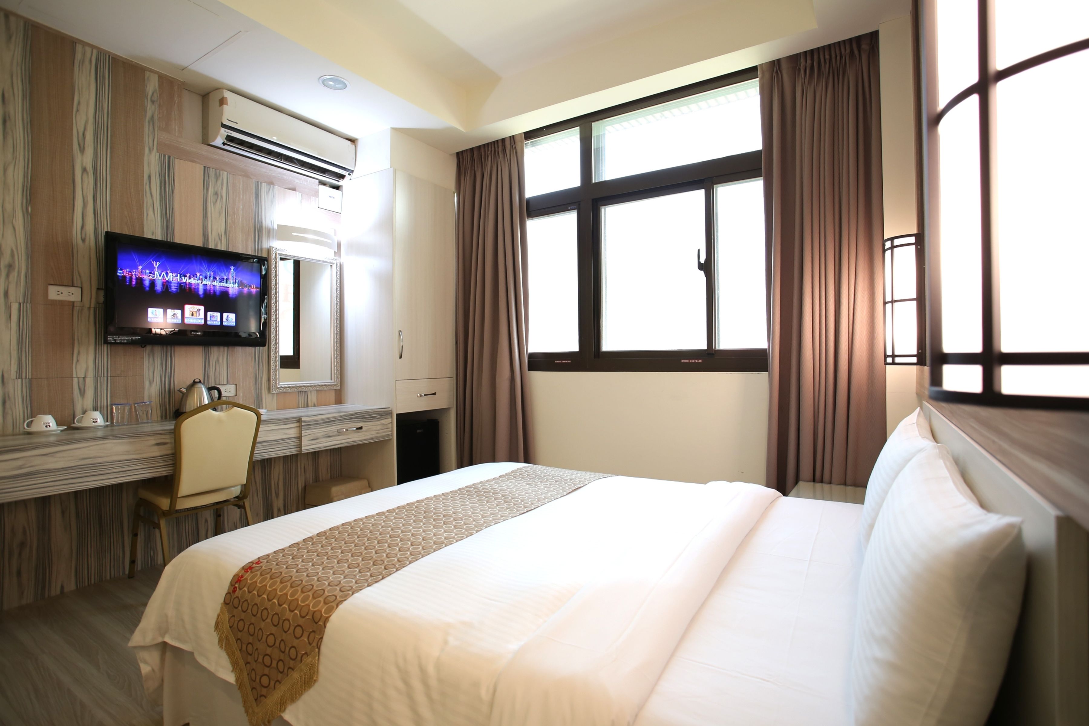 A hotel room with a large bed, a desk with a flat-screen TV, a chair, and a window with curtains.