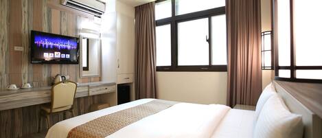 A hotel room with a large bed, a desk with a flat-screen TV, a chair, and a window with curtains.