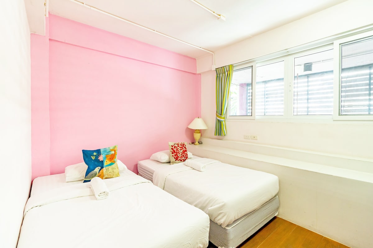 A room with two beds, a window with blinds, a lamp, and a pink wall.
