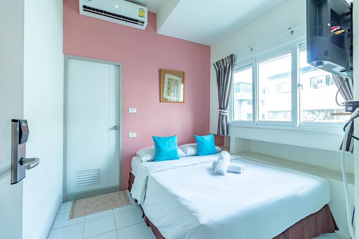 A hotel room with a bed, a door, an air conditioning unit, a window with curtains, and a wall-mounted television.