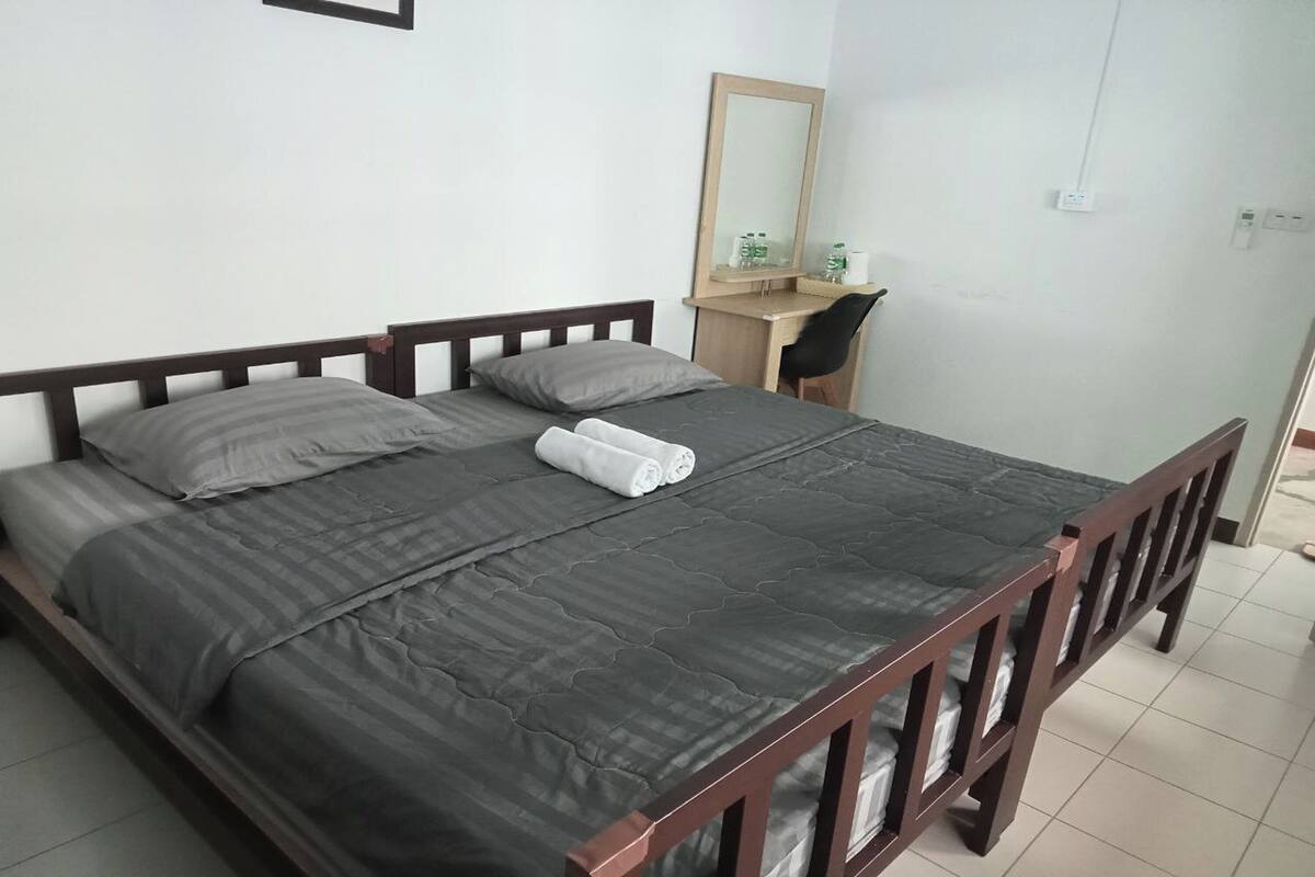A single bed with a dark gray quilt, two pillows, and a rolled white towel. A wooden desk with a chair is against the wall, and a mirror hangs above it.