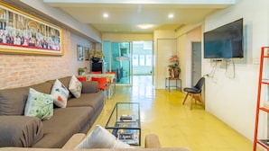 A living room with a sofa, coffee table, and a TV. There is a painting on the wall and a door leading to another room. - Sleep Dee Hostel (Bangkok)
