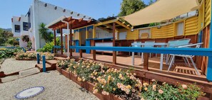 A covered outdoor seating area with blue railings and wooden tables, surrounded by flower beds and a building with yellow and blue exterior walls. - Te Aputa Lodge (Te Puru)