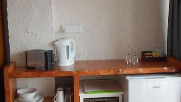 Cocina compacta con un refrigerador blanco, cafetera, tostadora y una computadora portátil sobre una encimera de madera. Hay cestas de mimbre y un estante con vajilla y un salero.