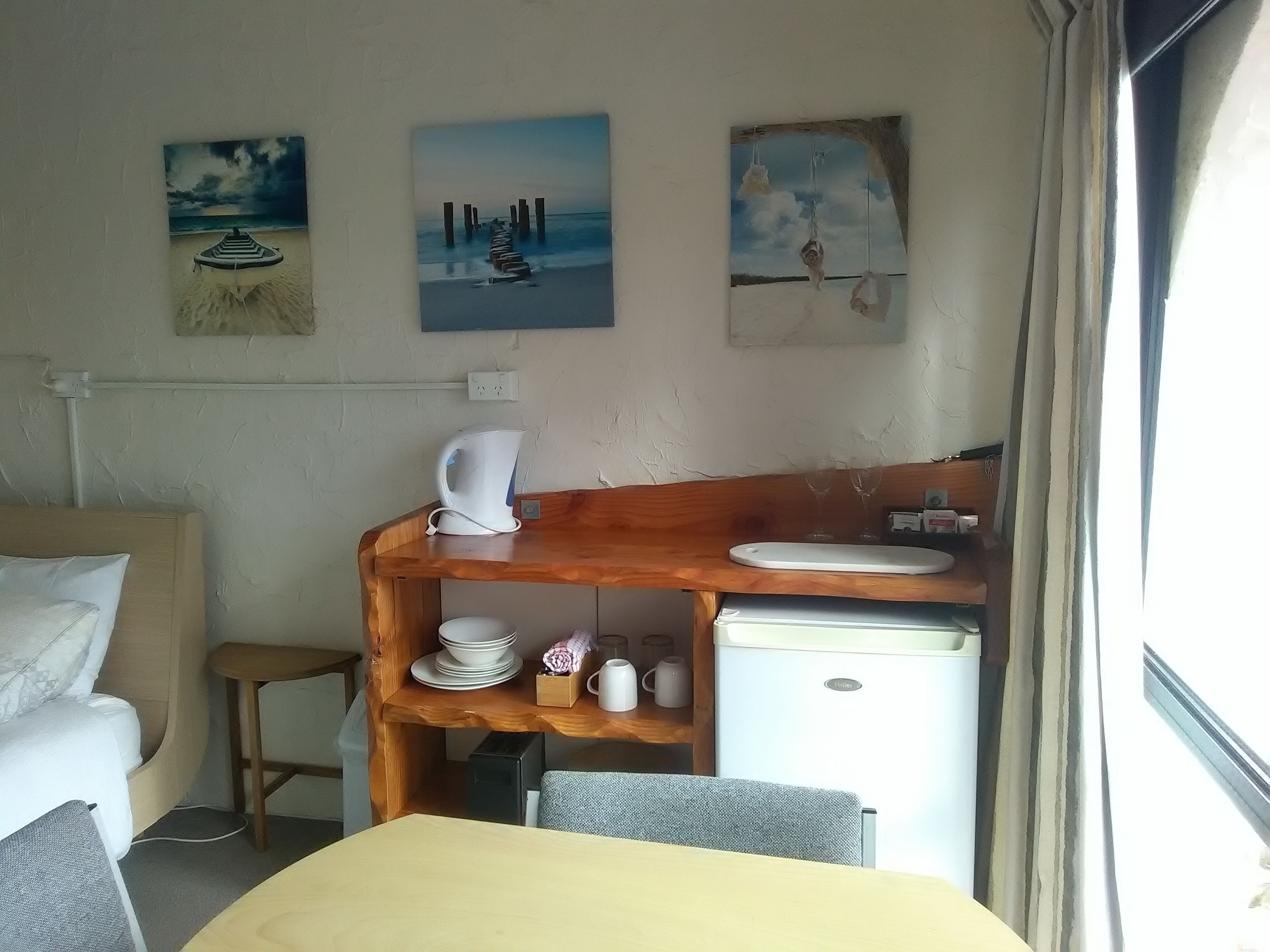 Una habitación compacta con cama, una mesita, un estante de madera con utensilios de cocina y un refrigerador. Hay tres cuadros en la pared.