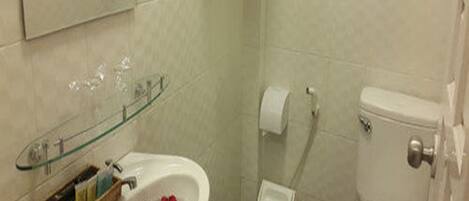 A bathroom with a glass shelf holding toiletries, a sink with rose petals, a toilet, and a trash bin.