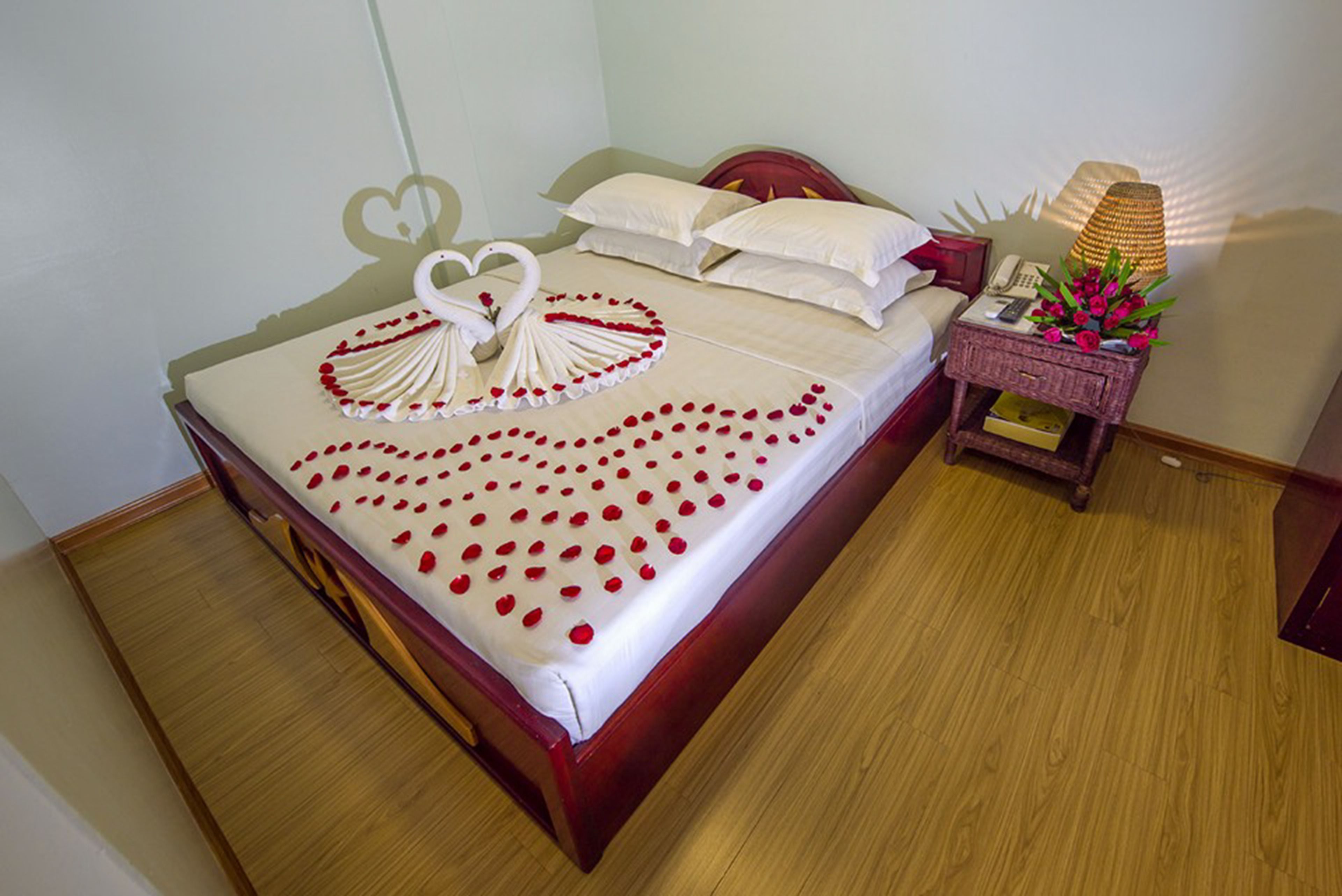 A neatly made bed with a heart-shaped decoration on the bedspread, a bedside table with a lamp, and a small box of tissues.