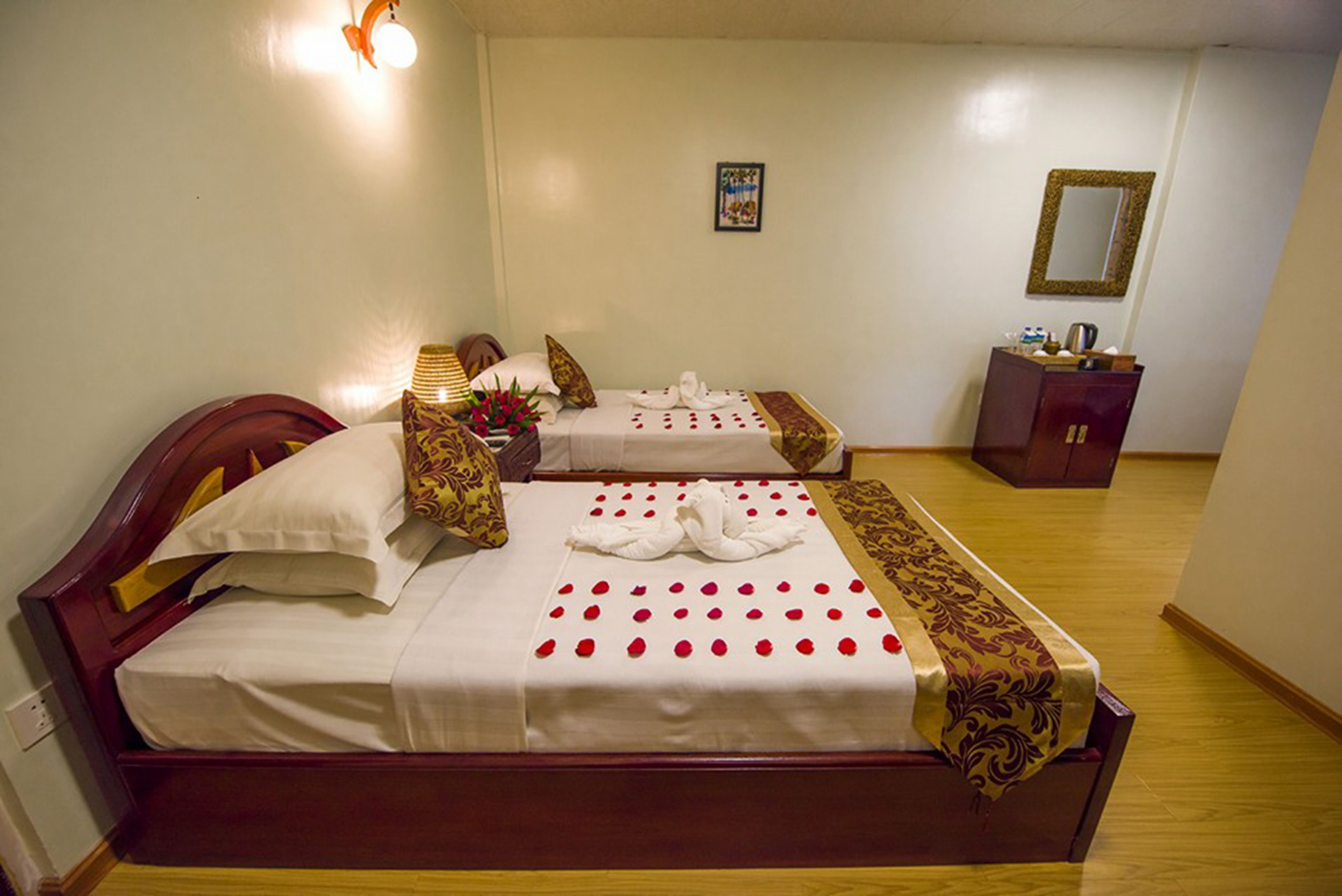 A hotel room with two beds, a wooden headboard, a bedside table with a lamp, a mirror, and a small cabinet.