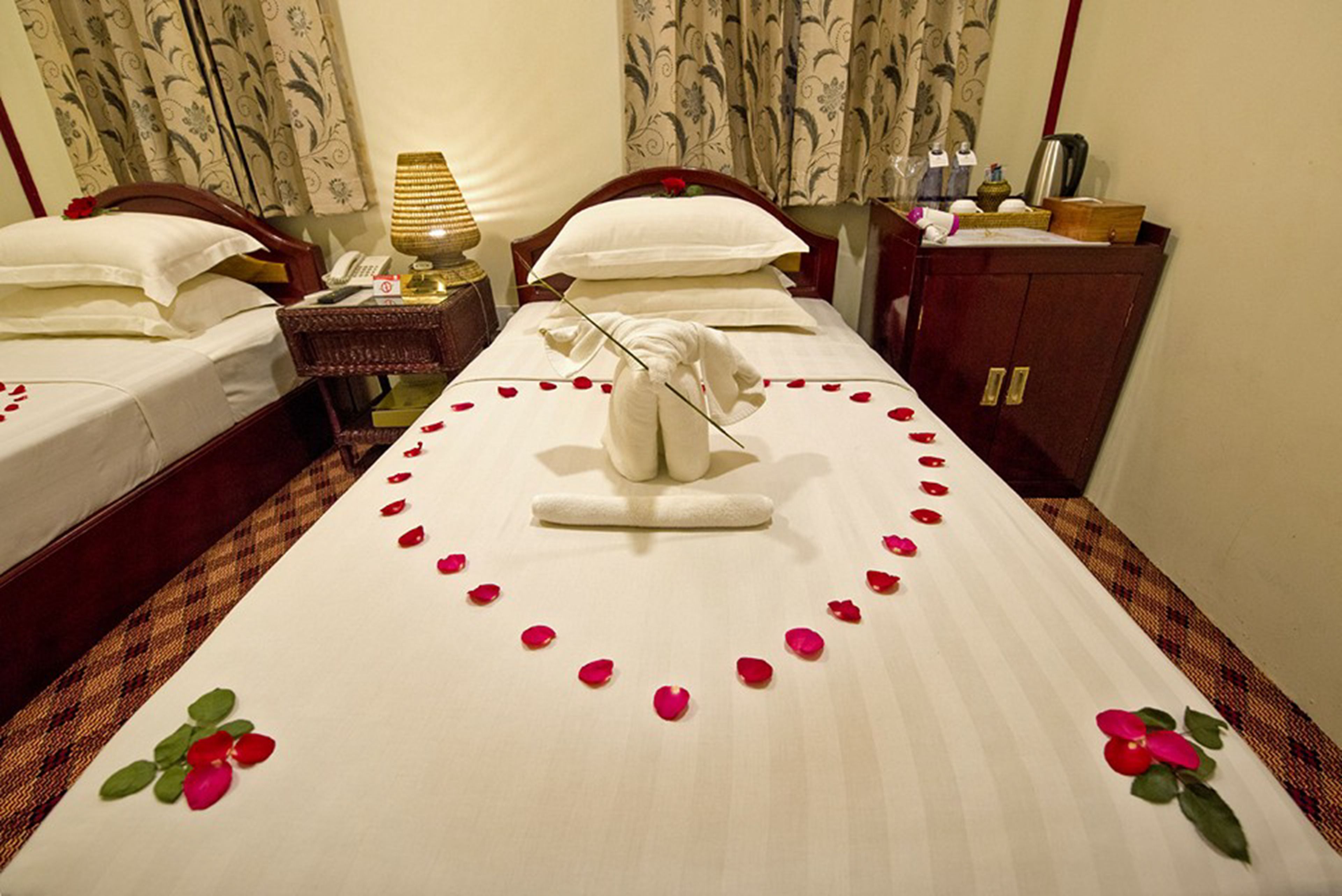 Quarto de hotel com duas camas, uma mesa de cabeceira com abajur, um armário de madeira e uma decoração de toalhas na cama.