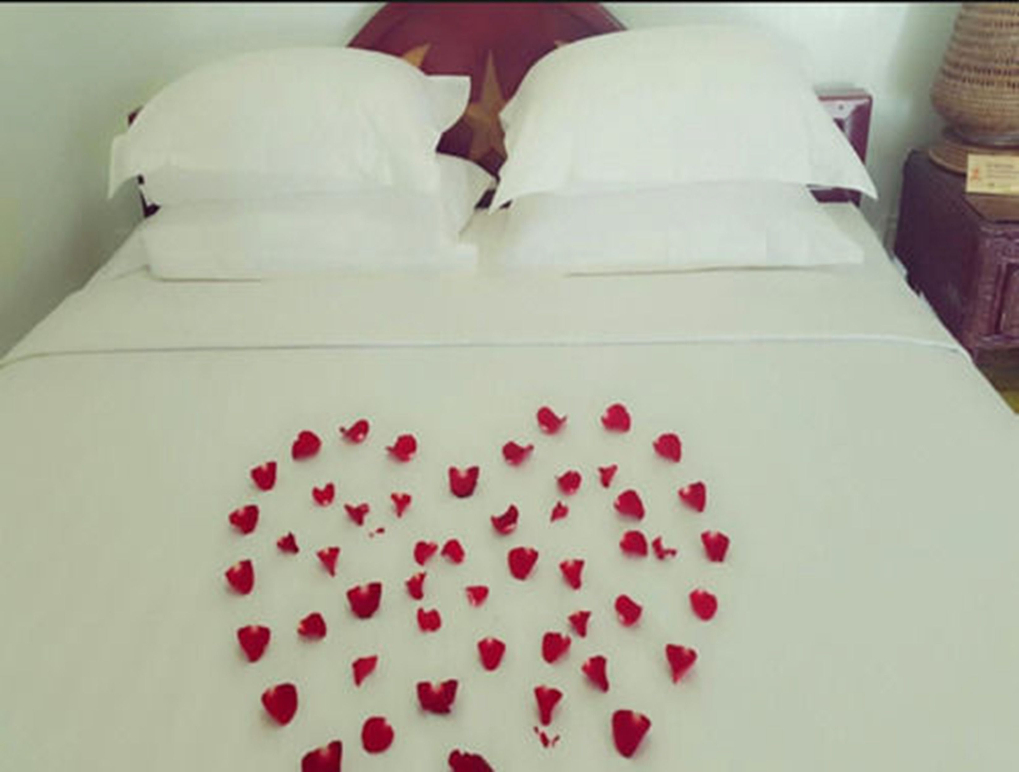 Una cama con sábanas blancas y un arreglo con pétalos de rosa roja en forma de corazón sobre el edredón.