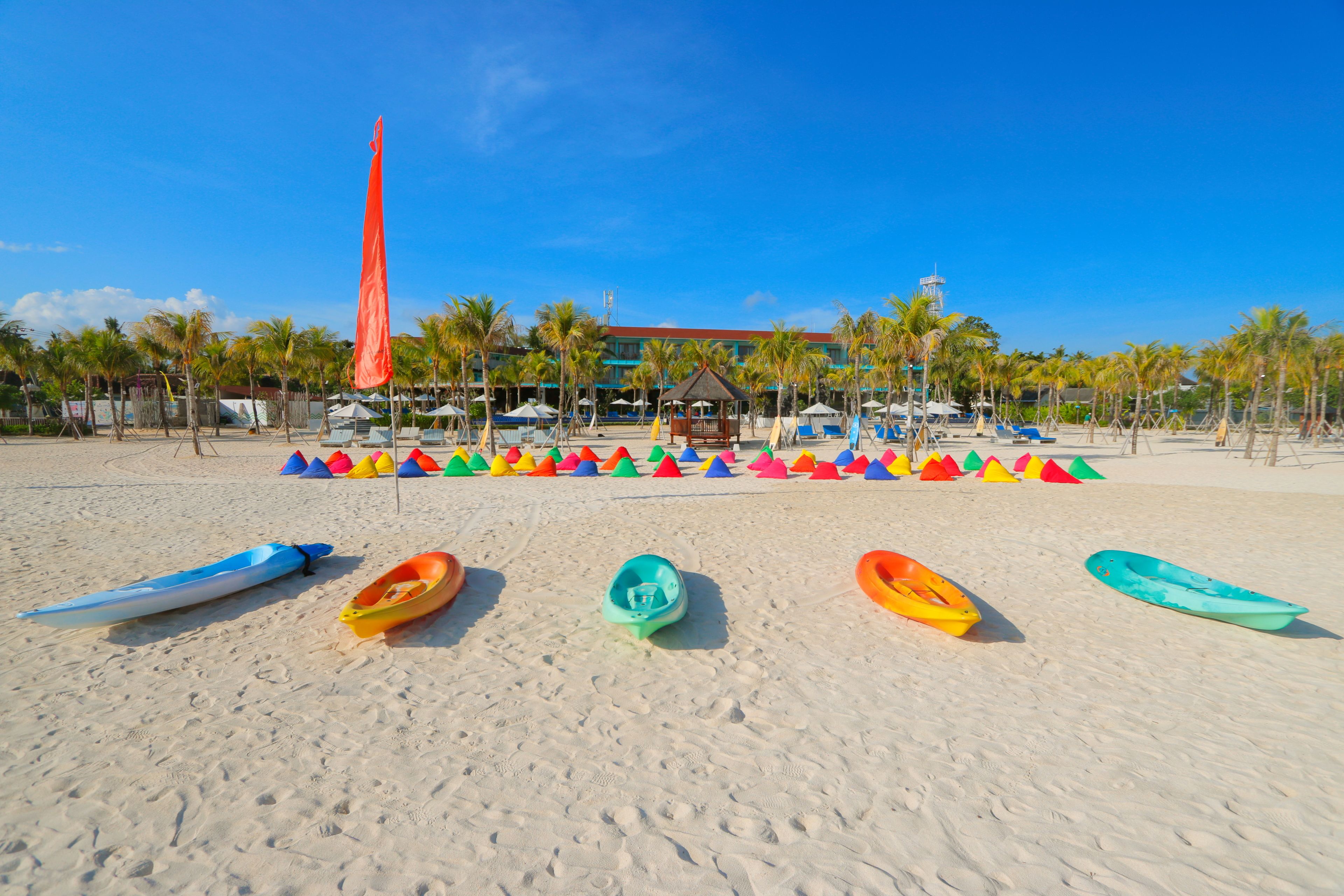 A beach with several colorful kayaks lined up in the foreground and a tall flagpole in the background.