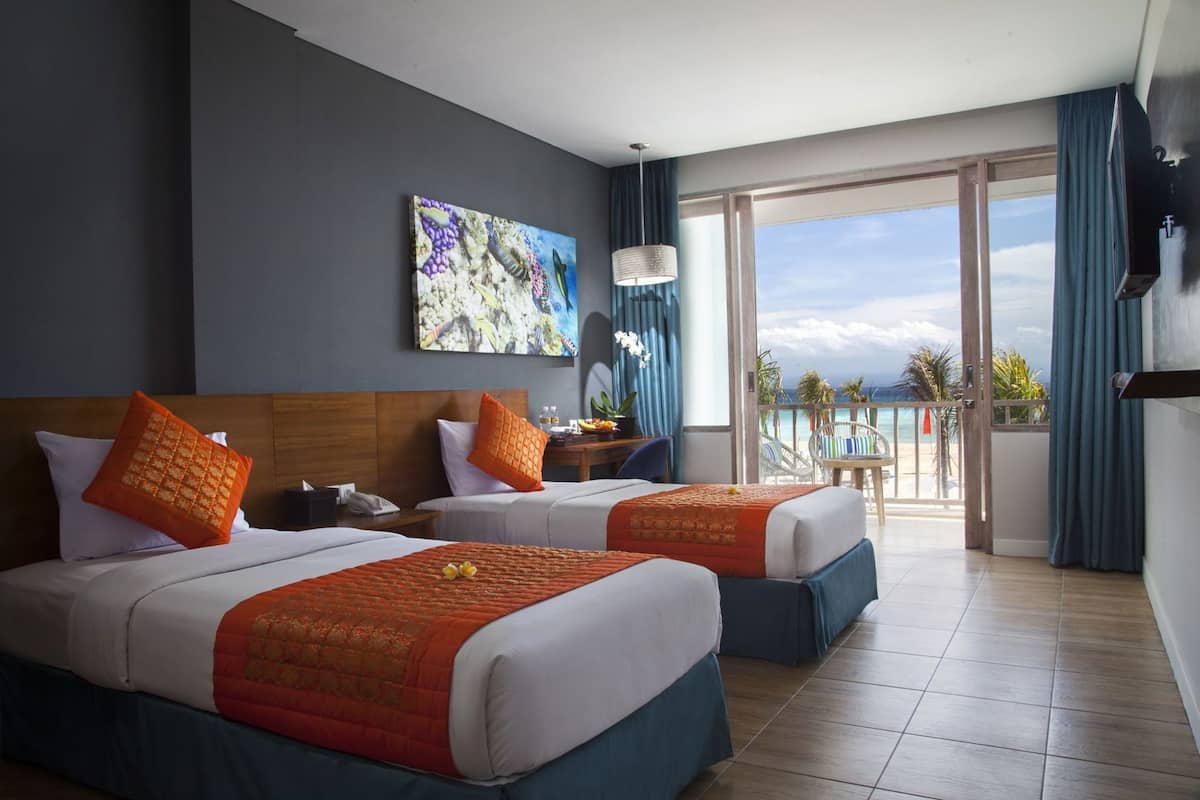 Ein Hotelzimmer mit zwei Betten, einem Gemälde, einem Schreibtisch mit Stuhl, einem Telefon und Blick auf den Ozean.
