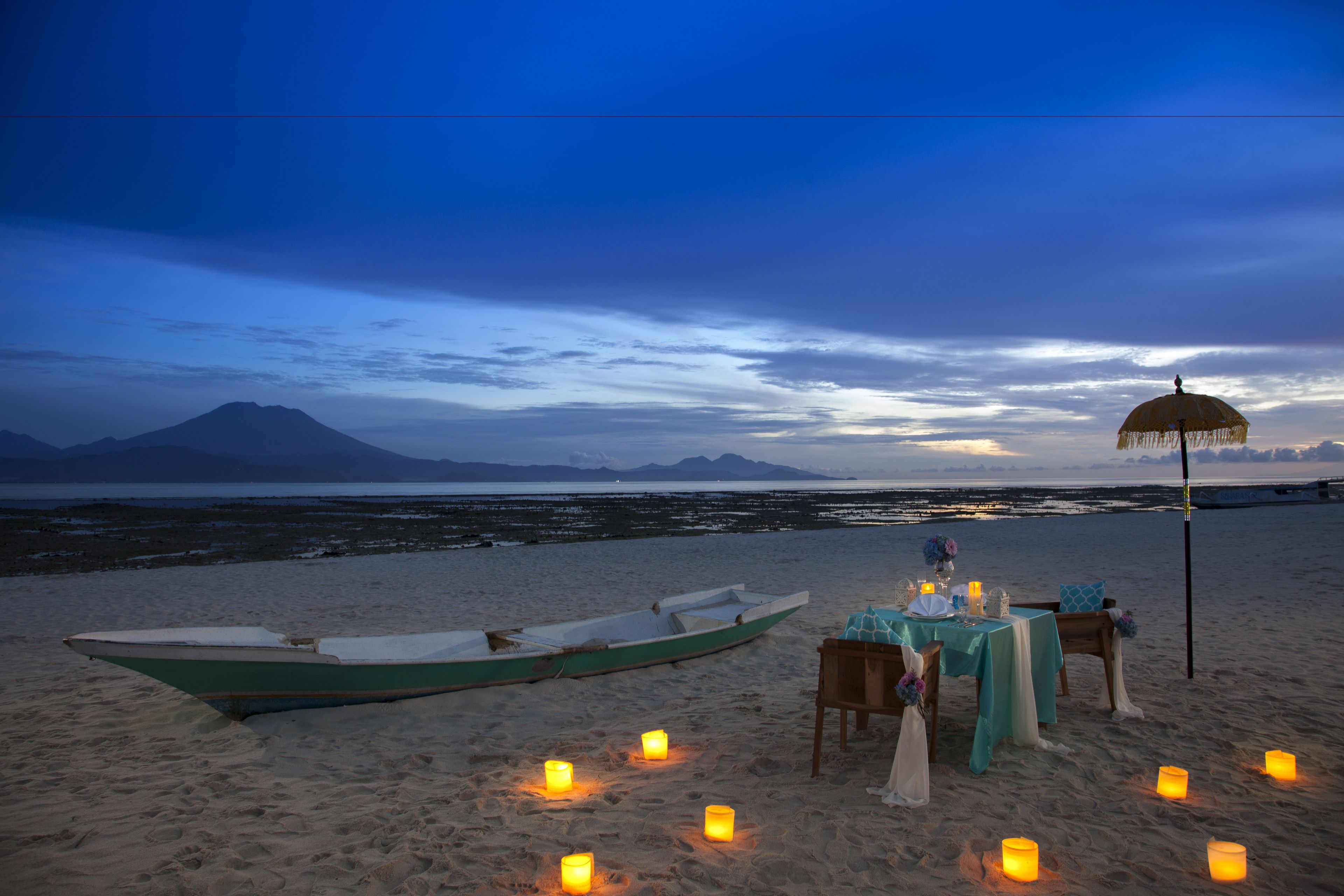 A beachside table set with candles, a boat, and a parasol.