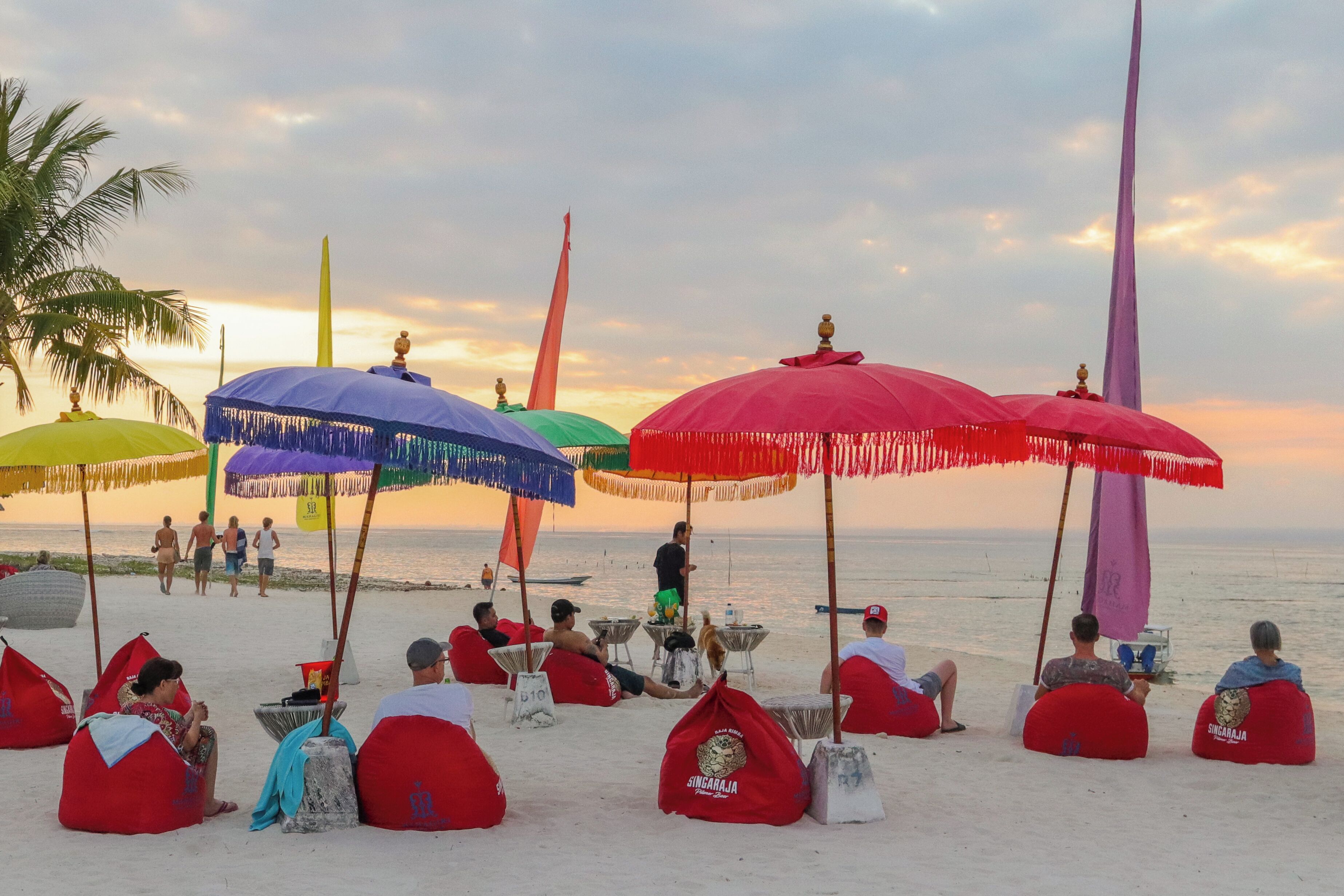 A beach with colorful umbrellas and bean bags, people relaxing, and a sunset in the background.