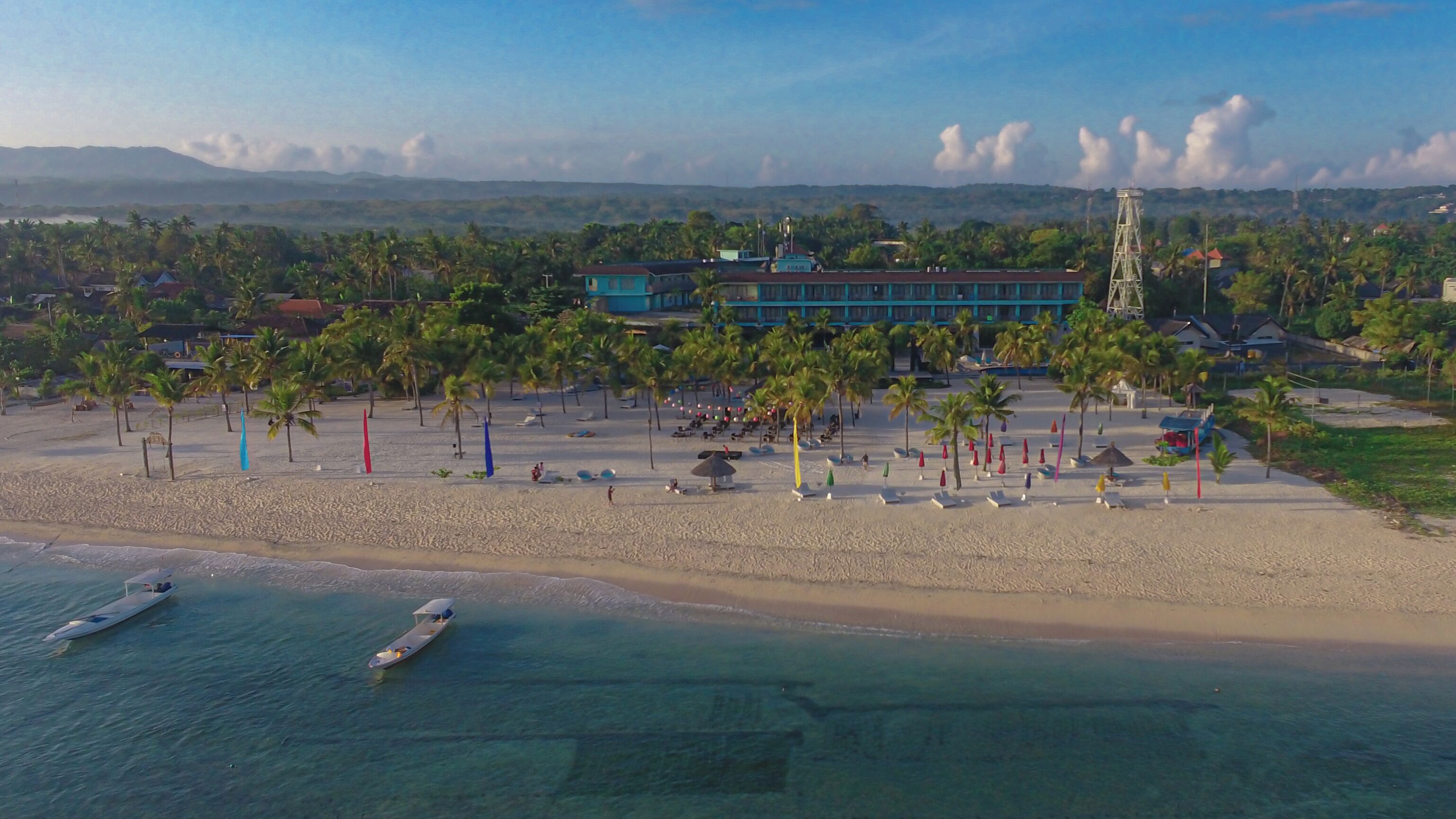 A beach with clear turquoise waters, a few boats, and a building with a blue roof in the background.