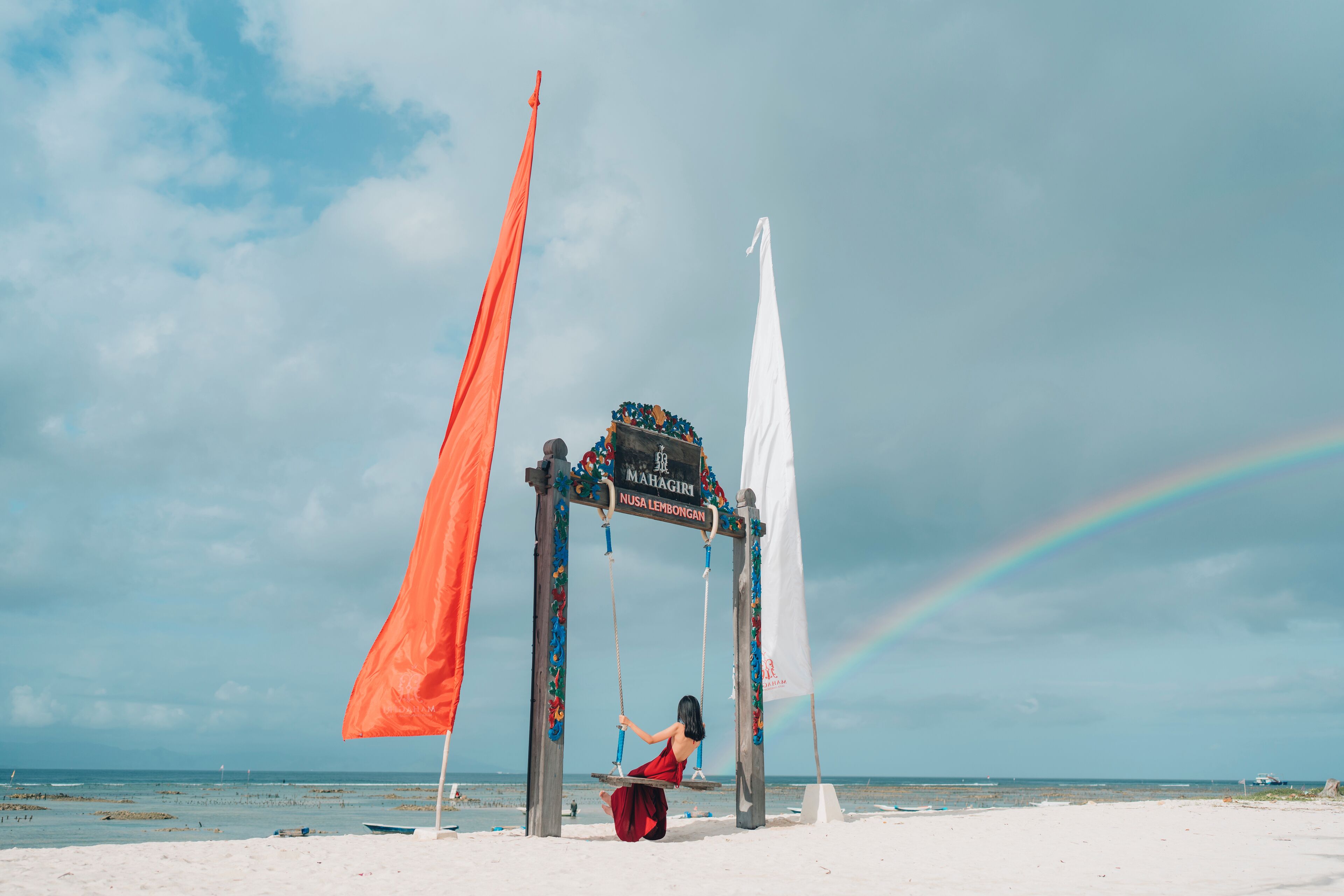 A beach scene with a swing set and a sign that reads "MAHAGIRI NUSA LEMBONGAN."