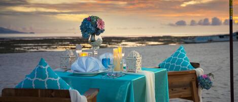 A beachside table set with a teal tablecloth, white napkins, and a vase of flowers.