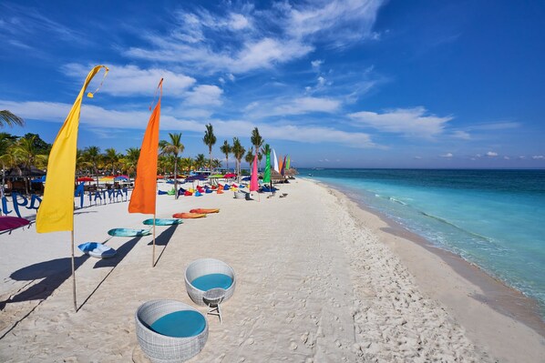 A beach with clear blue water, white sand, and colorful umbrellas and flags.