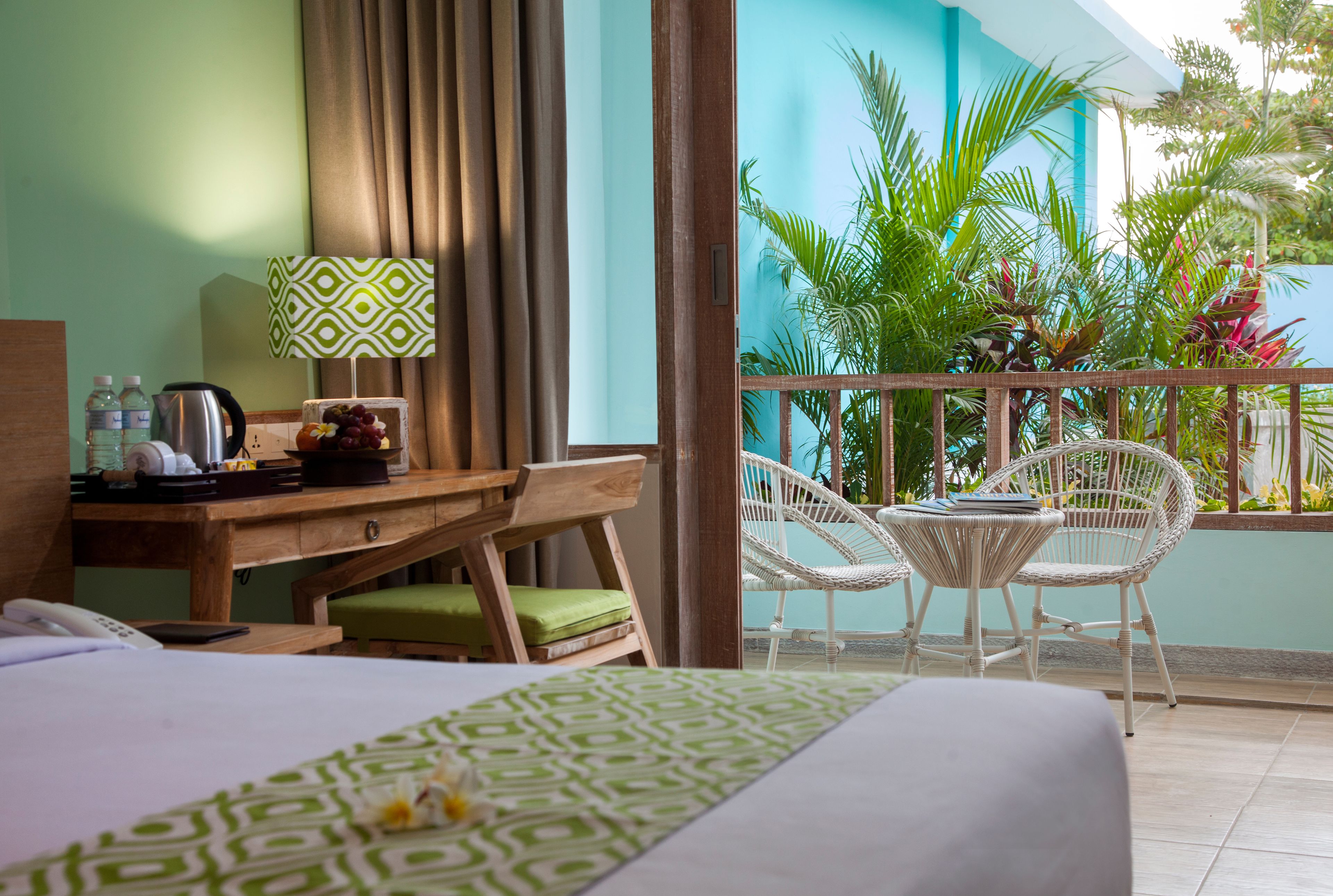 A hotel room with a bed, a desk with a lamp, a chair, and a balcony with plants.