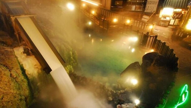 Un área de aguas termales con una cascada, rodeada de construcciones de piedra e iluminada con luz cálida.
