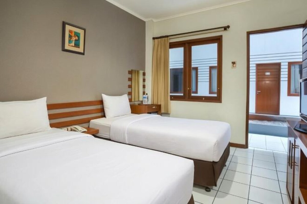 A hotel room with two beds, a bedside table with a lamp, a window with curtains, and a view of the outside.