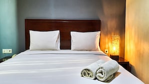 Un lit bien fait, avec du linge de lit blanc et des oreillers, une tête de lit en bois et une table de chevet fixée au mur, sur laquelle se trouve une lampe.