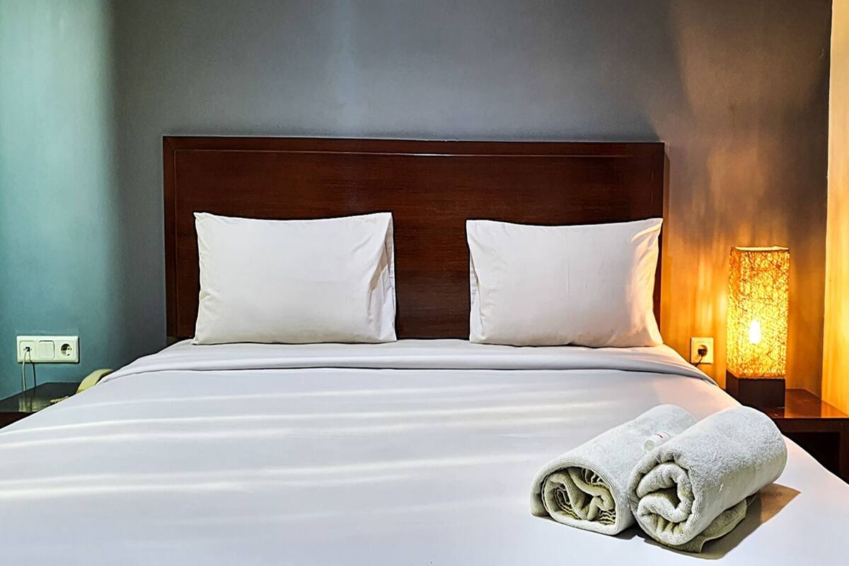 Een netjes opgemaakt bed met witte lakens en kussens, een houten hoofdbord, en een wandlampje op een nachtkastje.