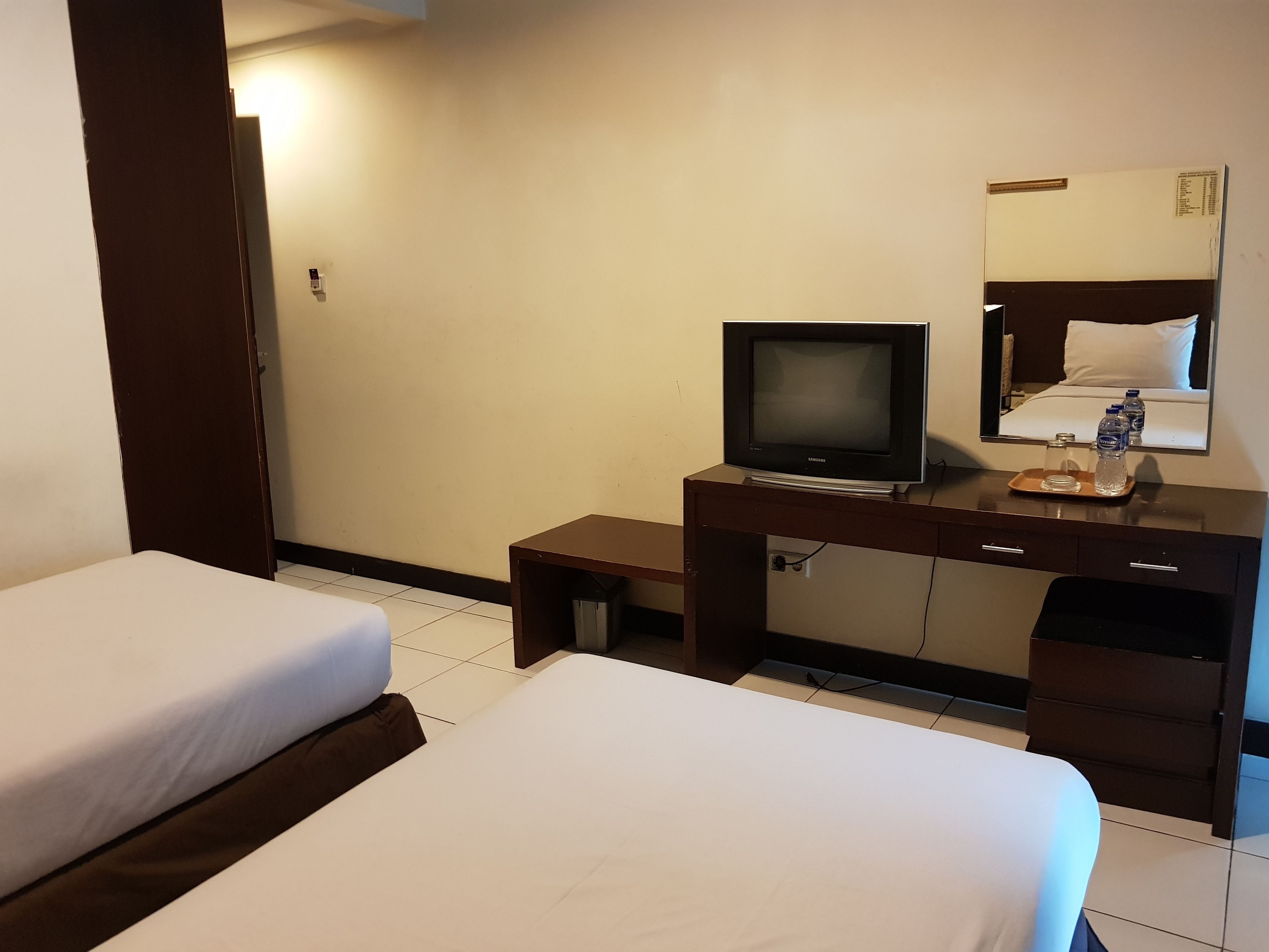 Habitación de hotel con dos camas, un televisor sobre un escritorio de madera, un espejo y una mesita con botellas de agua.