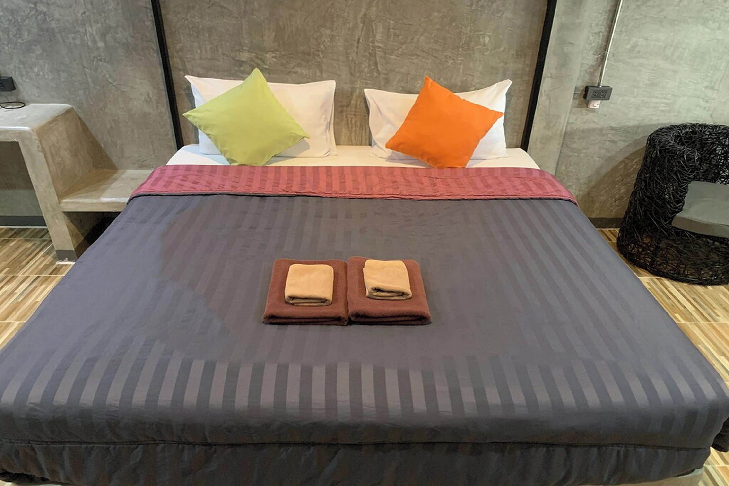 A neatly made bed with a dark bedspread, two folded towels, and two pillows in green and orange colors.