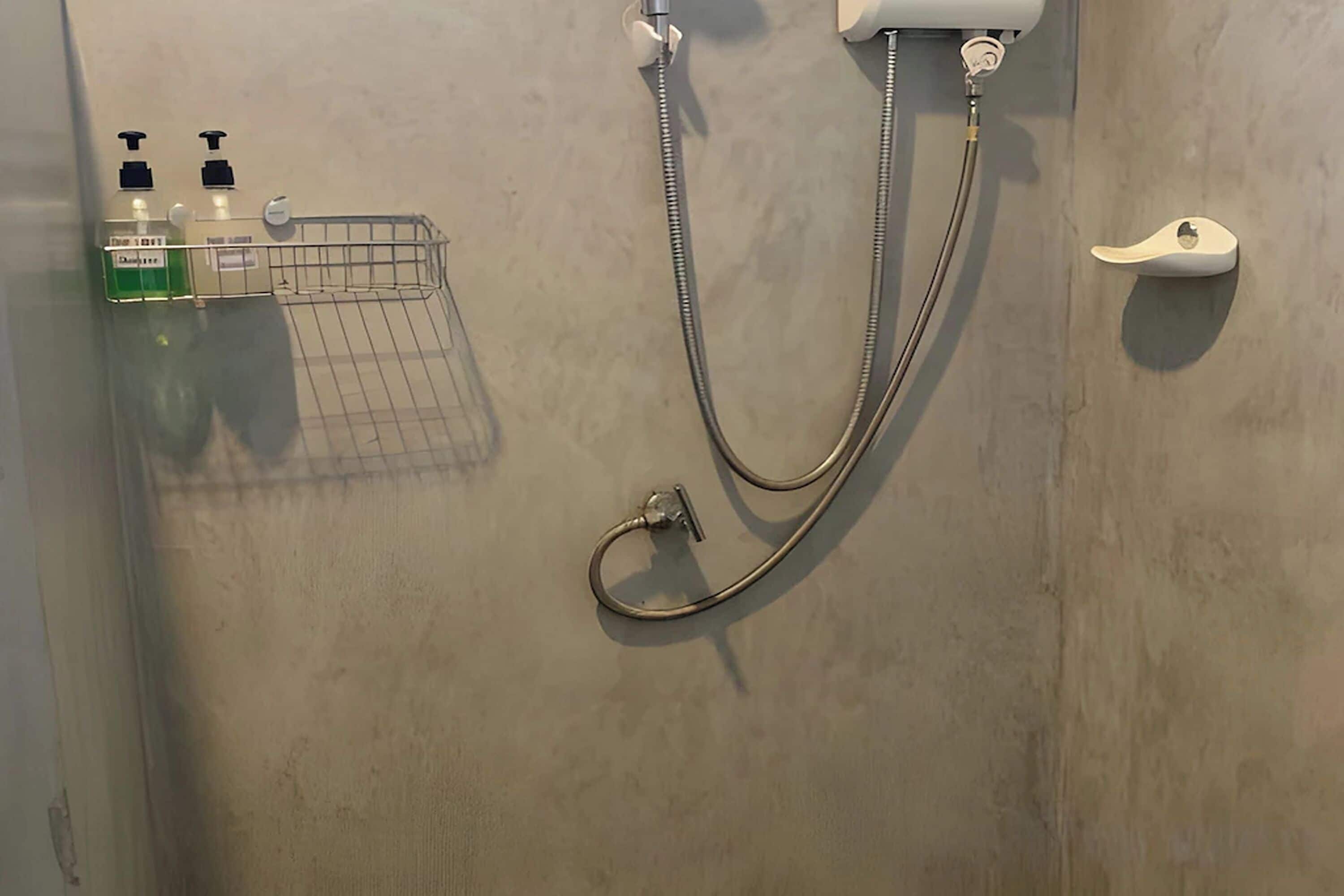 A shower with a handheld showerhead and a wall-mounted faucet. There is a soap dispenser and a small shelf with a towel.