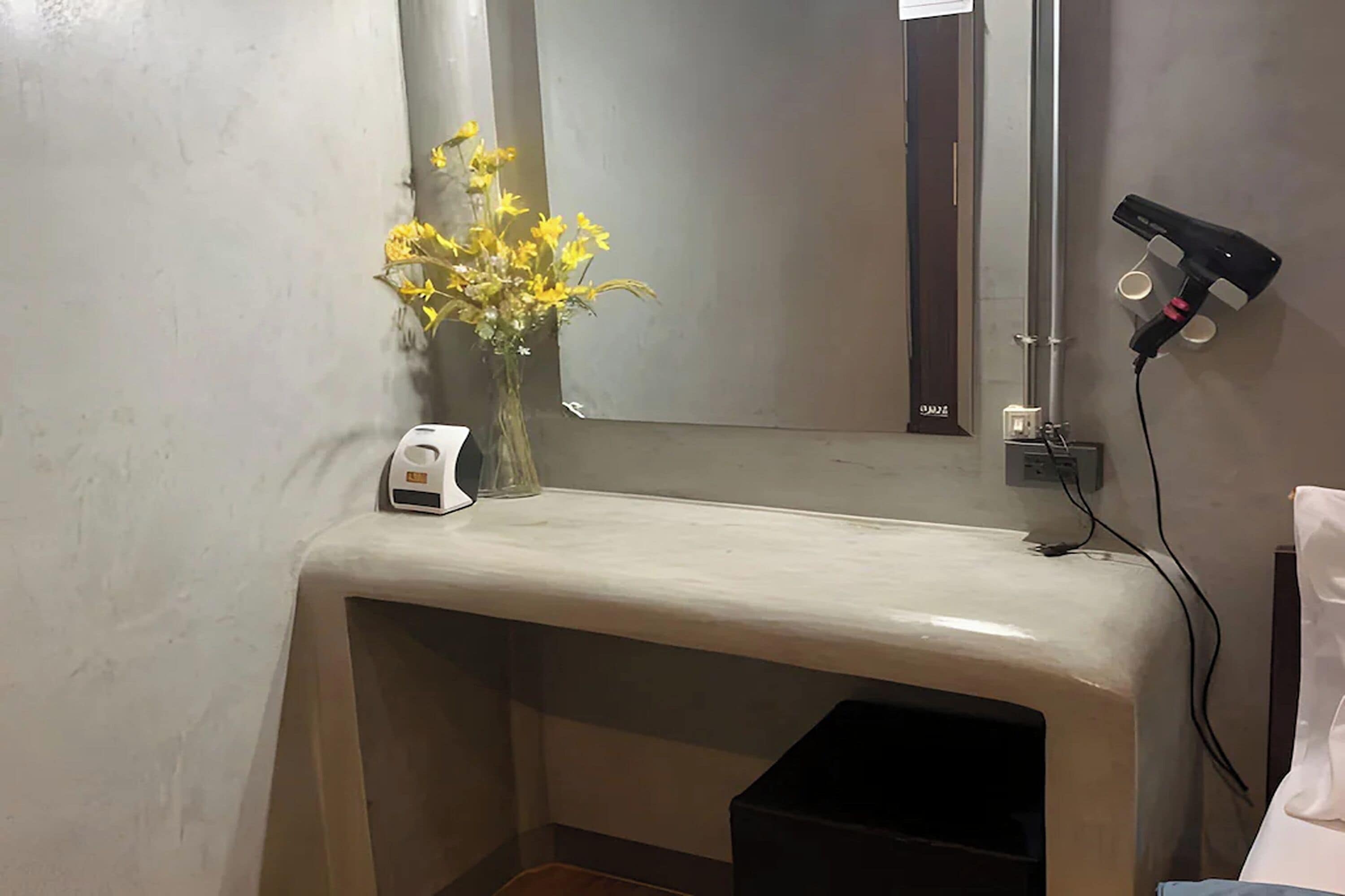 Un meuble-lavabo de salle de bain avec un miroir, un vase de fleurs jaunes, un sèche-cheveux et une petite poubelle.