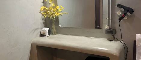 Un meuble-lavabo de salle de bain avec un miroir, un vase de fleurs jaunes, un sèche-cheveux et une petite poubelle.