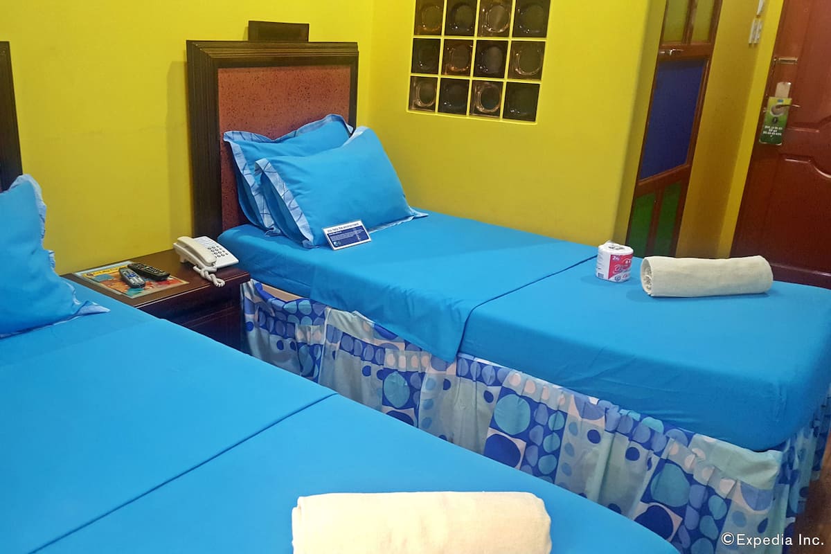 Quarto de hotel com duas camas, cada uma com roupa de cama e almofadas azuis, uma mesinha de cabeceira com telefone e controles remotos, e uma janela com vista para o exterior.