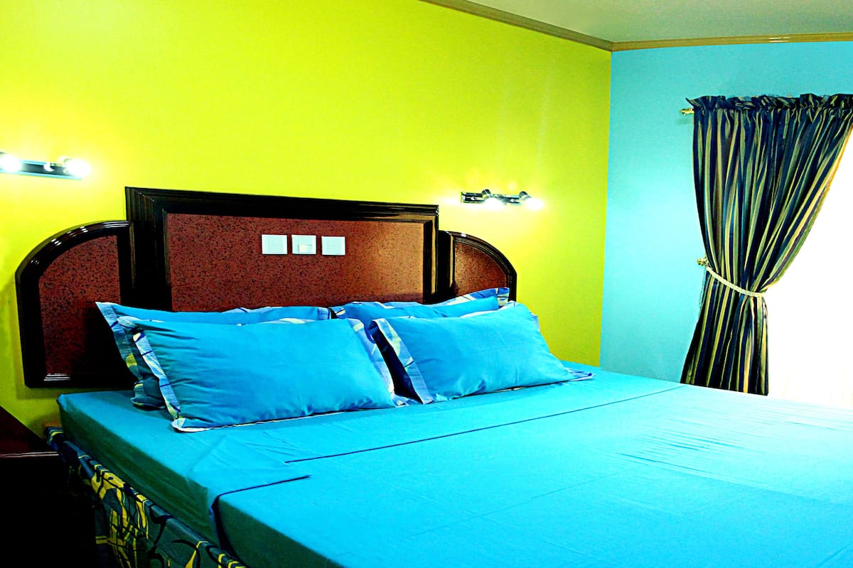 Cama com cabeceira, duas almofadas e colcha azul em quarto com paredes amarelas e janela com cortinas.