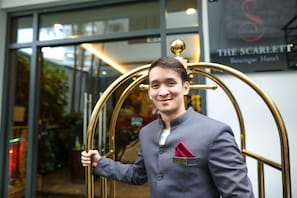 A man in a grey suit with a red pocket square standing in front of a hotel entrance with a sign reading "The Scarlett Boutique Hotel".