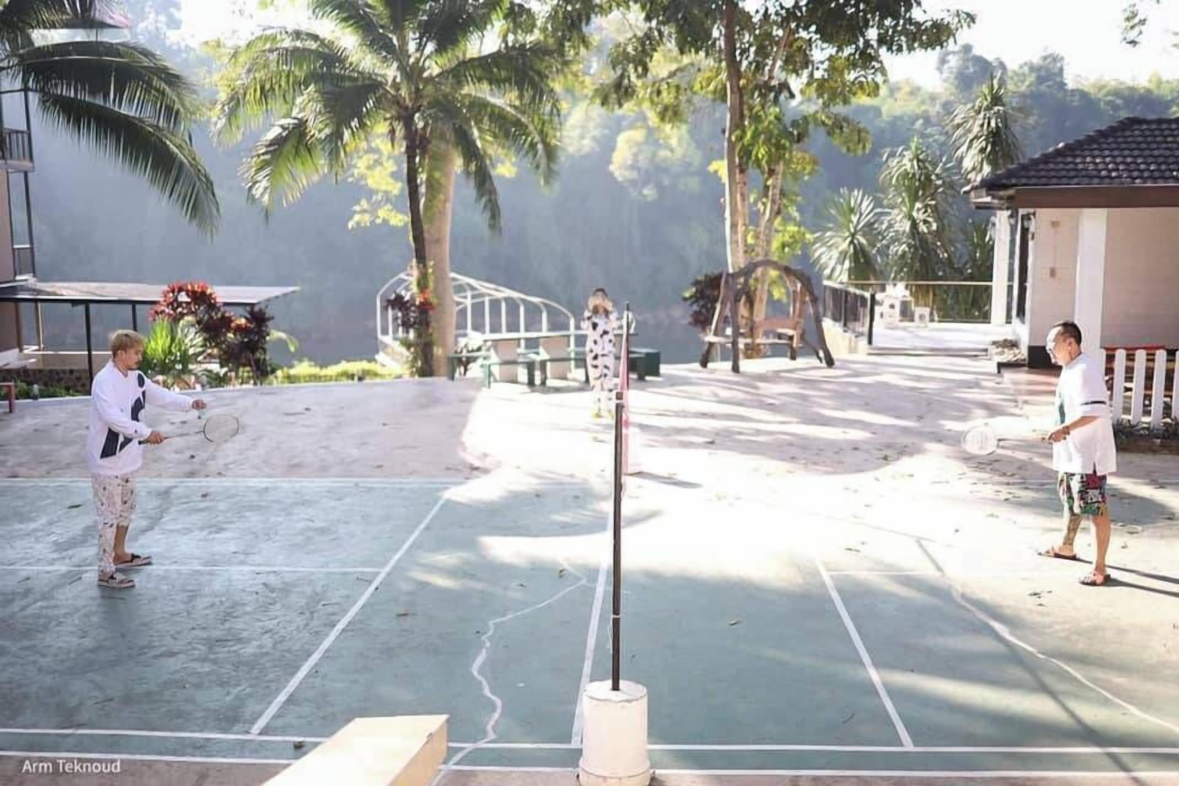 A tennis court with two players, a net, and a palm tree in the background.