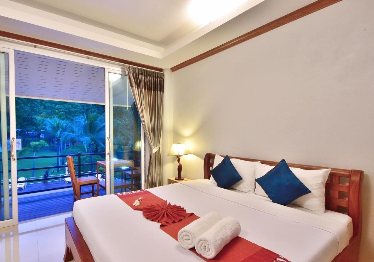 A hotel room with a bed, a nightstand, a lamp, a window with curtains, and a view of a pool and greenery.