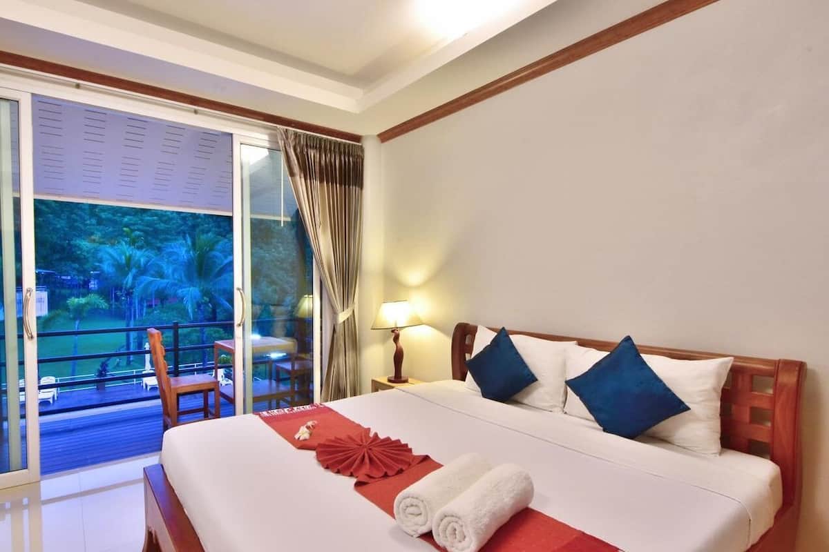 A hotel room with a bed, a nightstand, a lamp, a window with curtains, and a view of a pool and greenery.