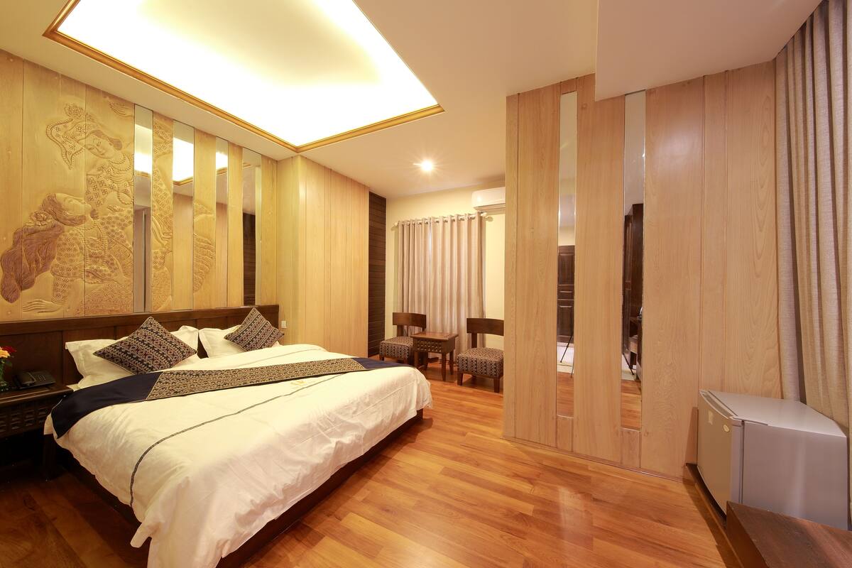 A hotel room with a large bed, wooden floor, a desk with a chair, a small table, and a refrigerator.