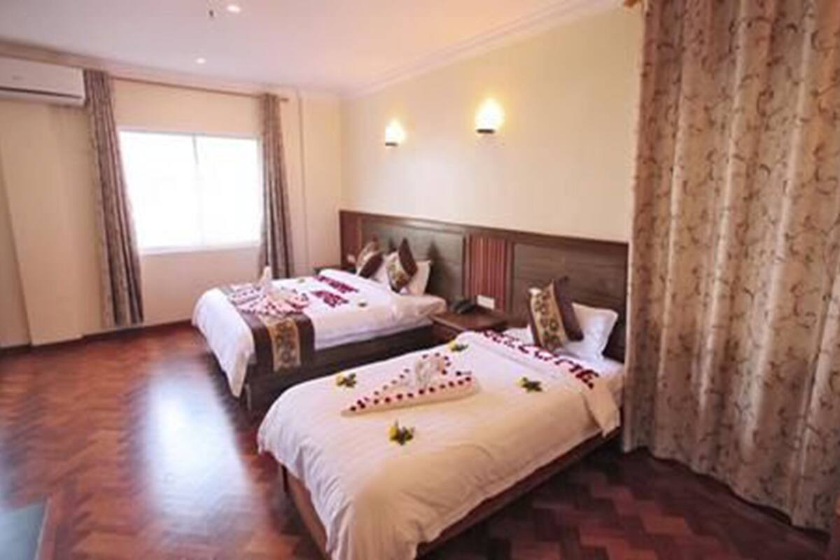 A hotel room with two beds, a wooden floor, a window with curtains, and a headboard with wall-mounted lights.