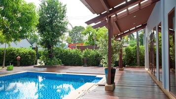 A swimming pool with a wooden deck, surrounded by lush greenery and a covered area with large windows.