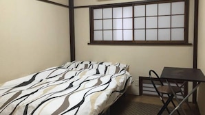 Una habitación pequeña con una cama, una mesa y una silla. Hay una ventana con diseño de cuadrícula y un aire acondicionado encima.